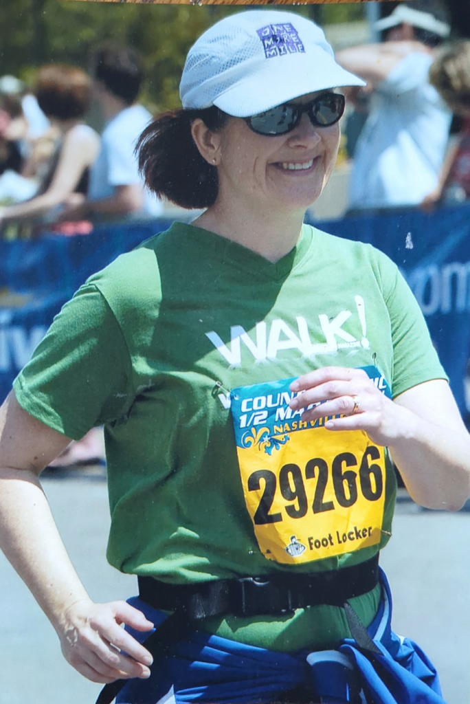 Cindi Leeman in a green shirt, white hate and yellow race bib walking the Country Music Half Marathon.