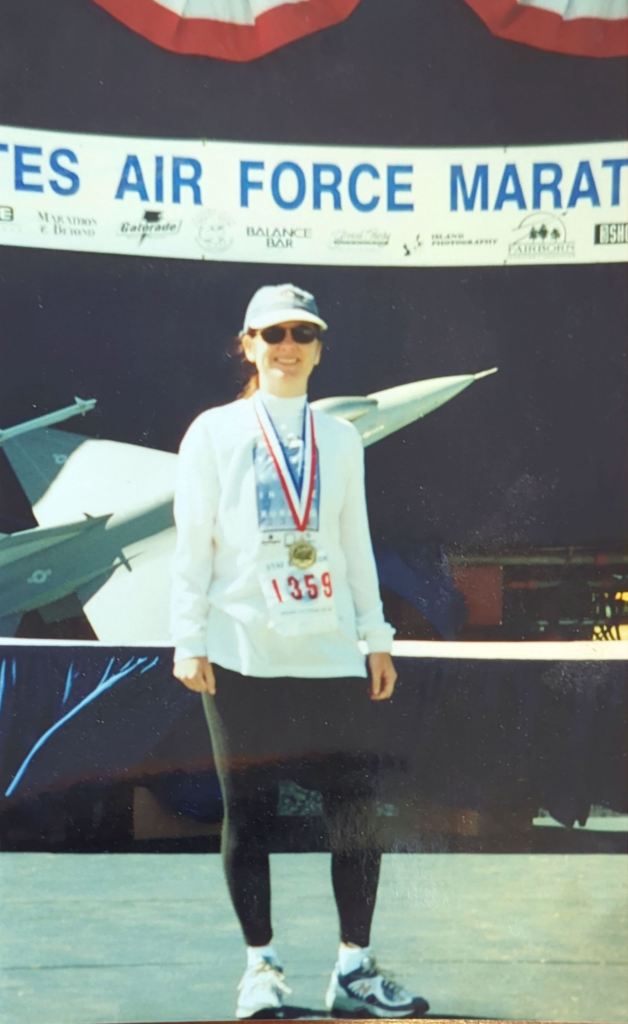 Cindi Leeman wearing a white shirt, blue hat, and race medal from the Air Force Marthon.
